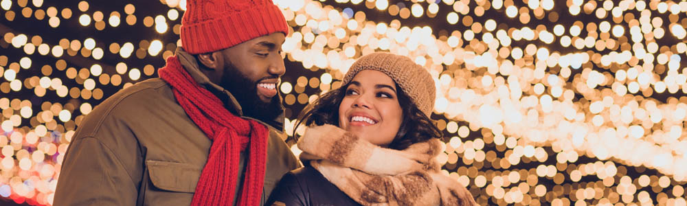 Couple dressed in their winter gear (with hats & scarves), smiling at each other with twinkling Christmas lights in the background